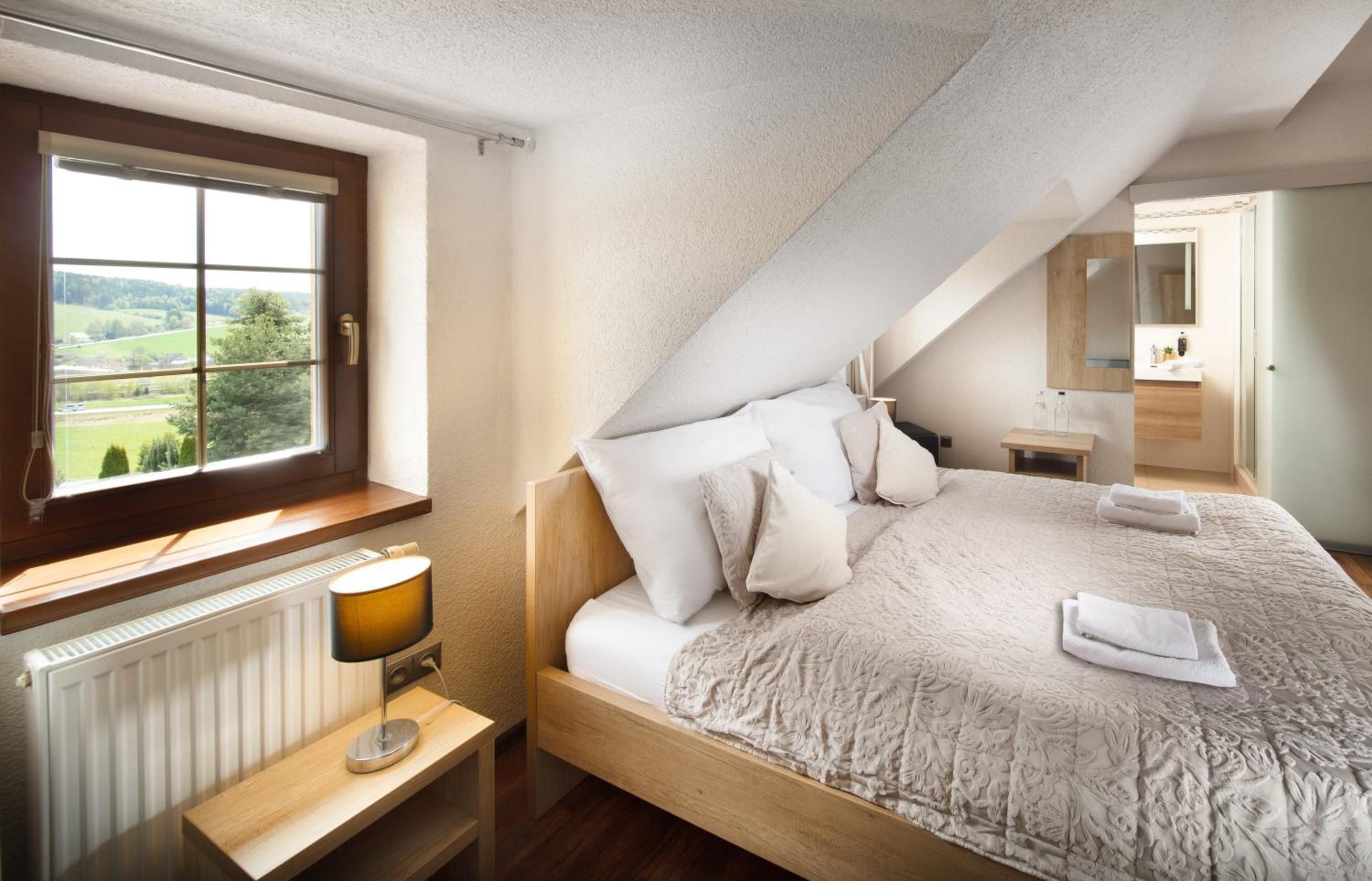A bedroom with a large bed, a window with a view, a bedside table with a lamp, and a bathroom visible in the background.