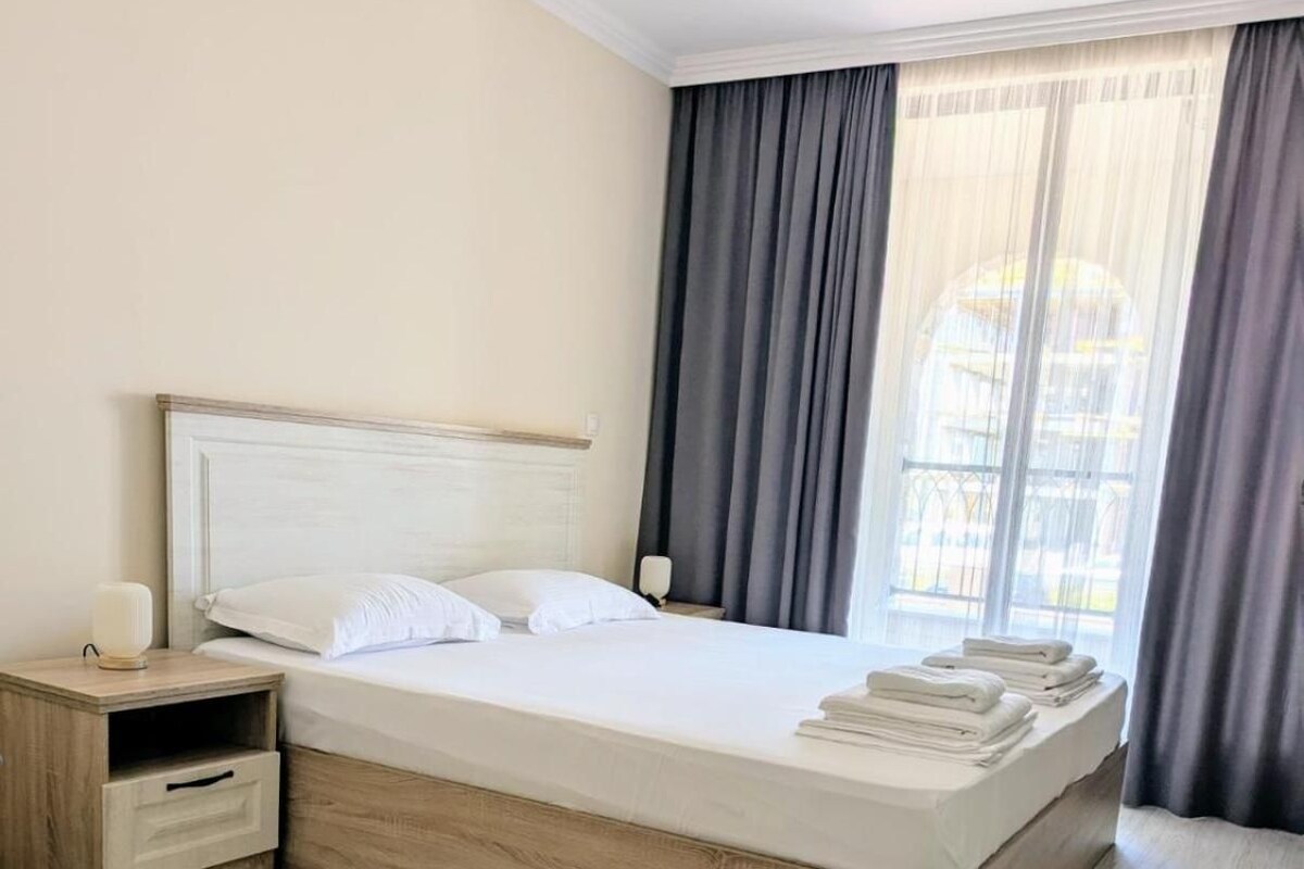 A hotel room with a large bed, a bedside table, a nightstand, and a window with curtains.