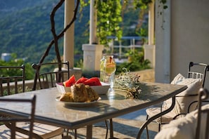A table set with a glass of white wine, watermelon slices, and a plate of bread, with a view of greenery in the background.