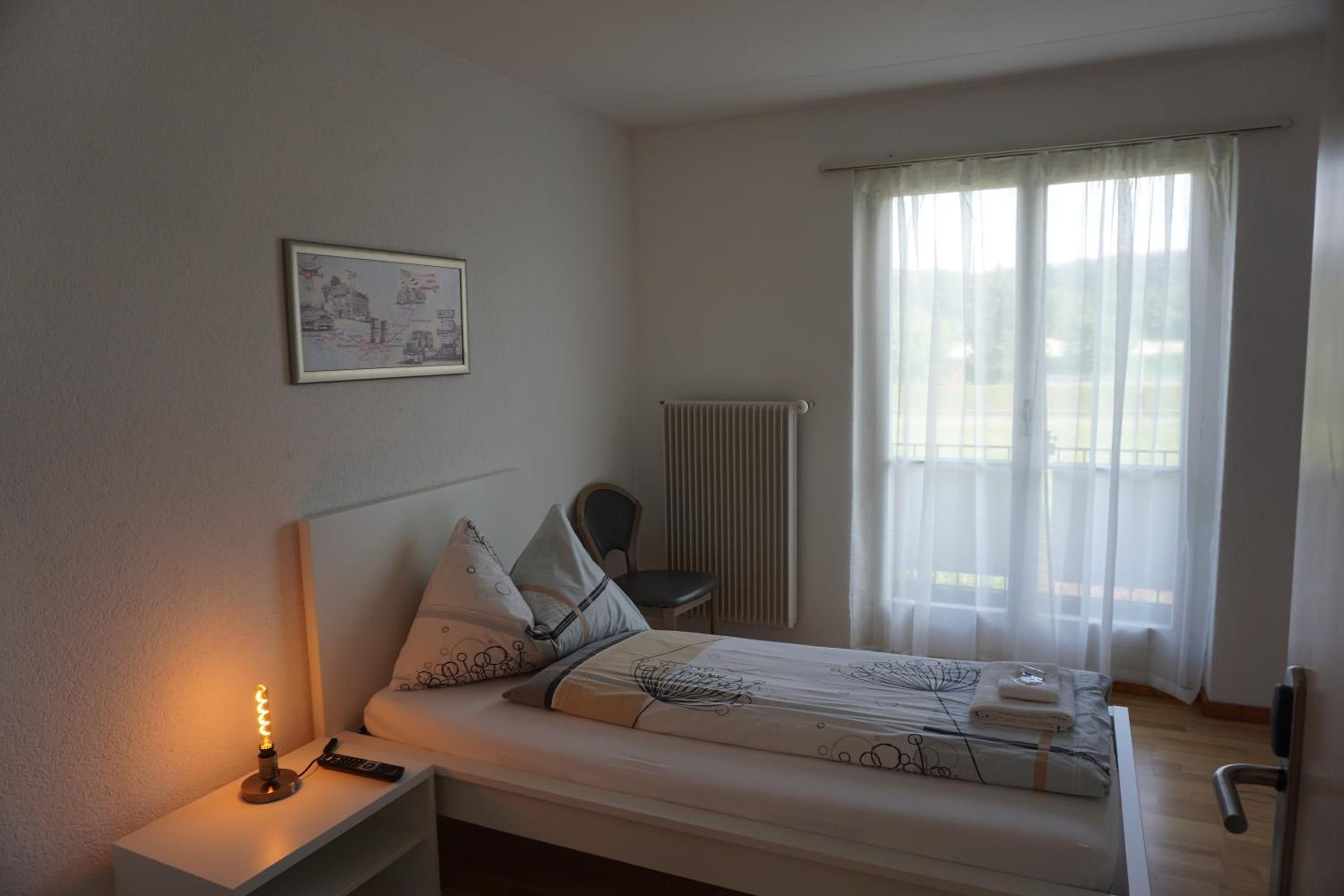 A single-bed room with a bedside table, a lamp, a chair, a radiator, and a window with curtains.