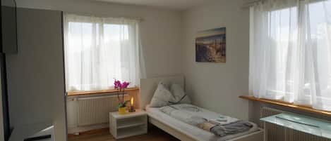 A compact room with a single bed, a small bedside table, a radiator, and a window with sheer curtains.