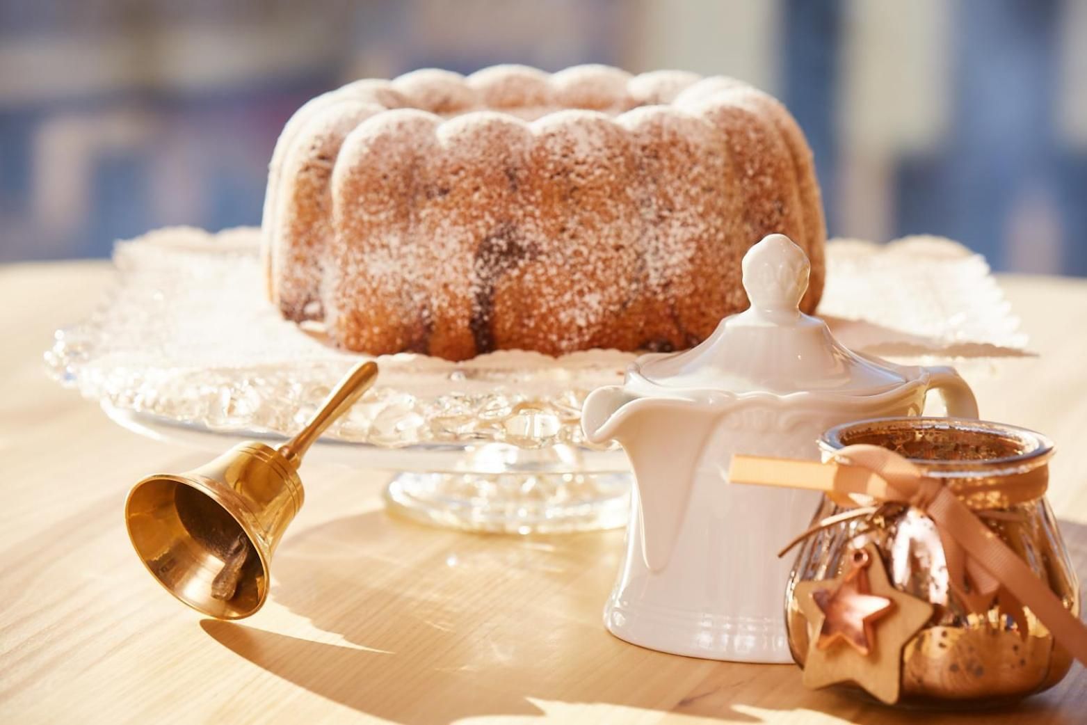 Un pastel bundt sobre un soporte de vidrio, una jarrón blanco y un frasco con tapa en forma de estrella sobre una mesa de madera.
