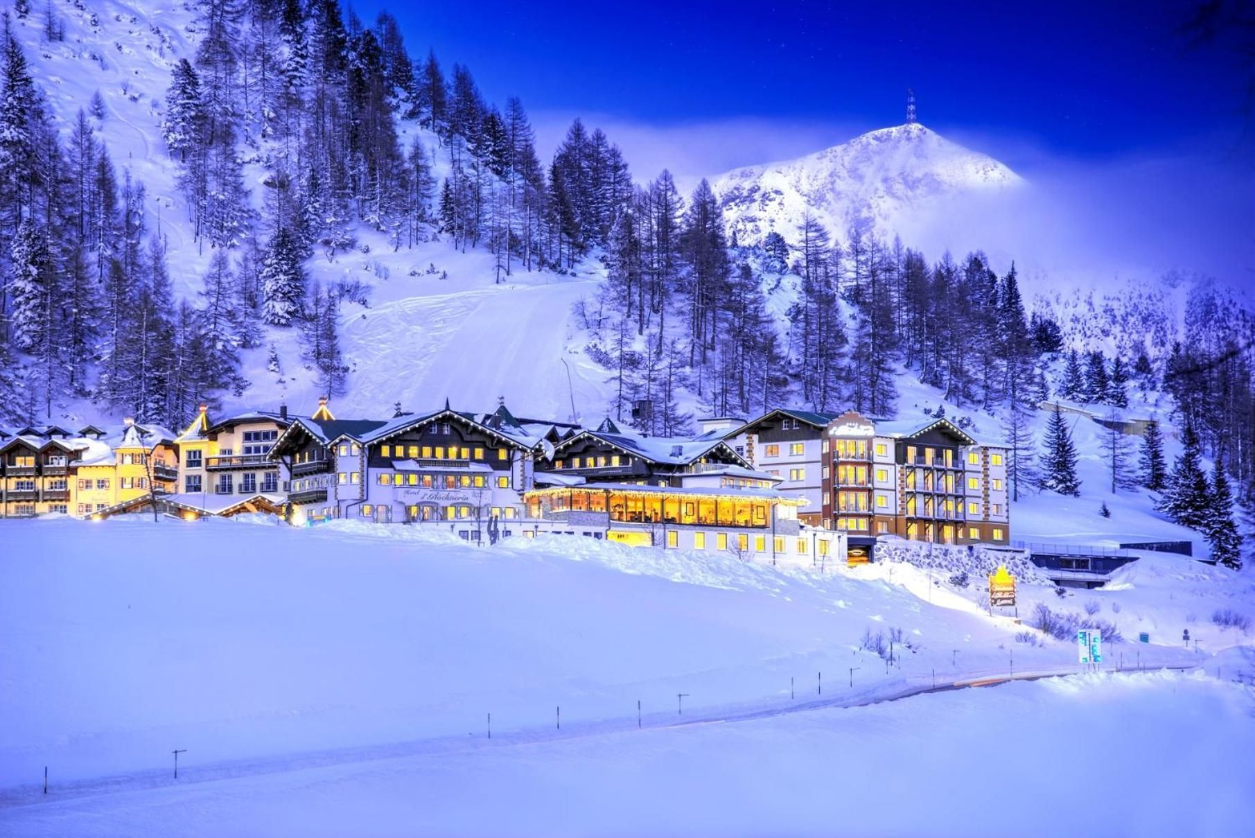 Estância de montanha coberta de neve, com vários edifícios, incluindo um hotel com varandas e um teleférico visível ao fundo.