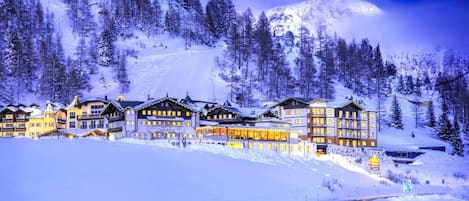 A snow-covered mountain resort with multiple buildings, including a hotel with balconies and a ski lift visible in the background.