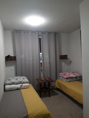 A small, compact room with two single beds, a small table, and a shelf.