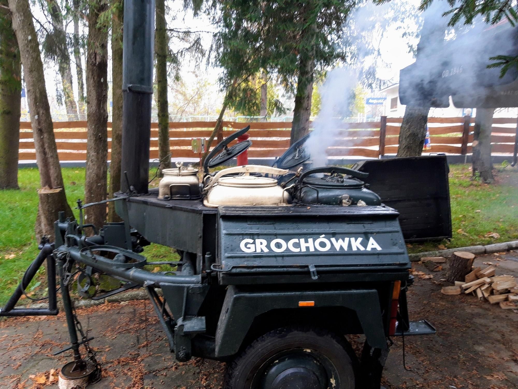 A portable cooking device labeled 'GROCHOWKA' with a chimney emitting smoke, set up outdoors with trees and a fence in the background.