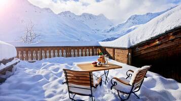 Une table en bois placée à l’extérieur, entourée de chaises, dans un paysage enneigé, avec des montagnes en arrière-plan.