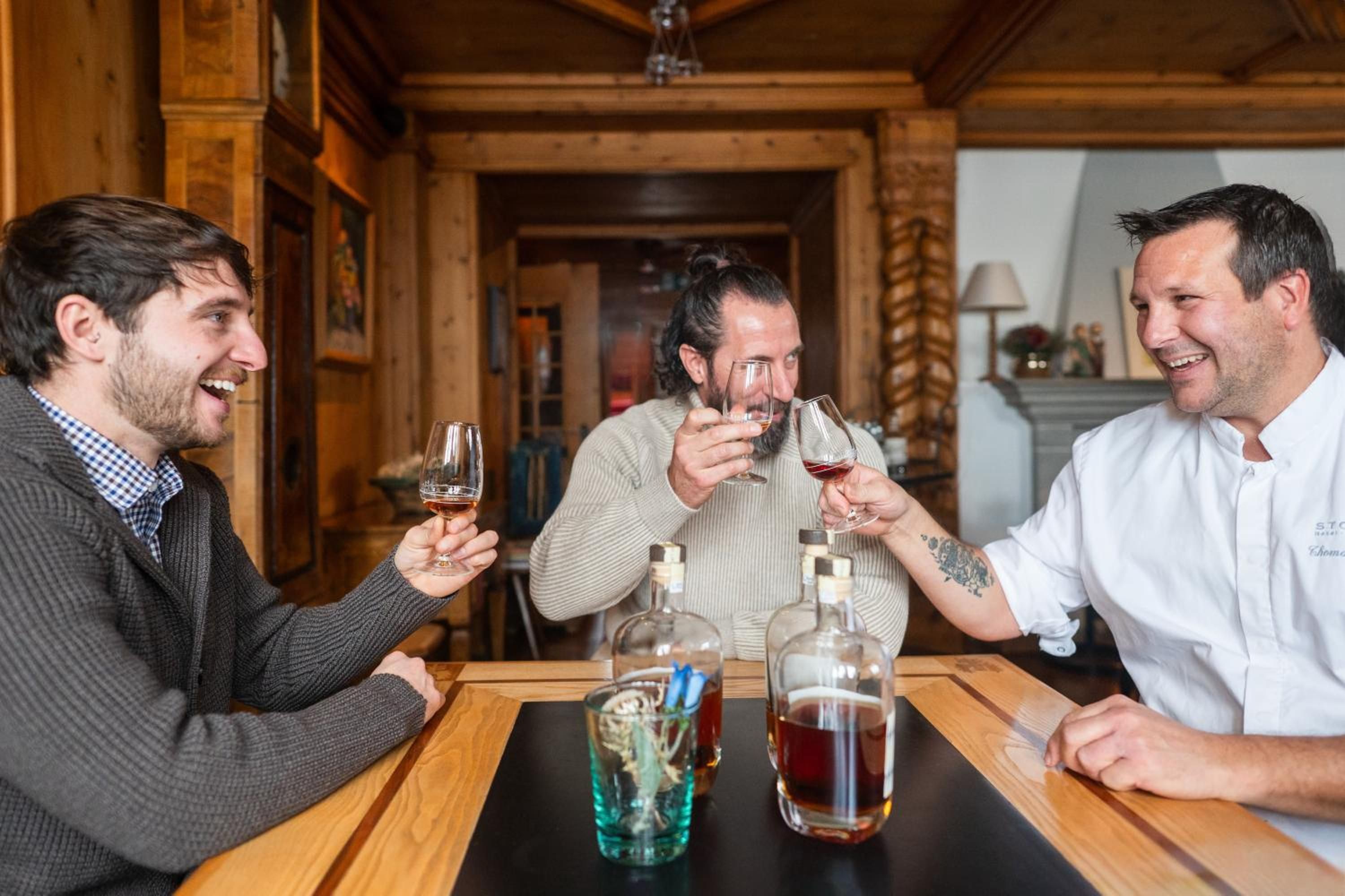 Tres personas sentadas en una mesa de madera con vasos, una botella y un recipiente de vidrio.