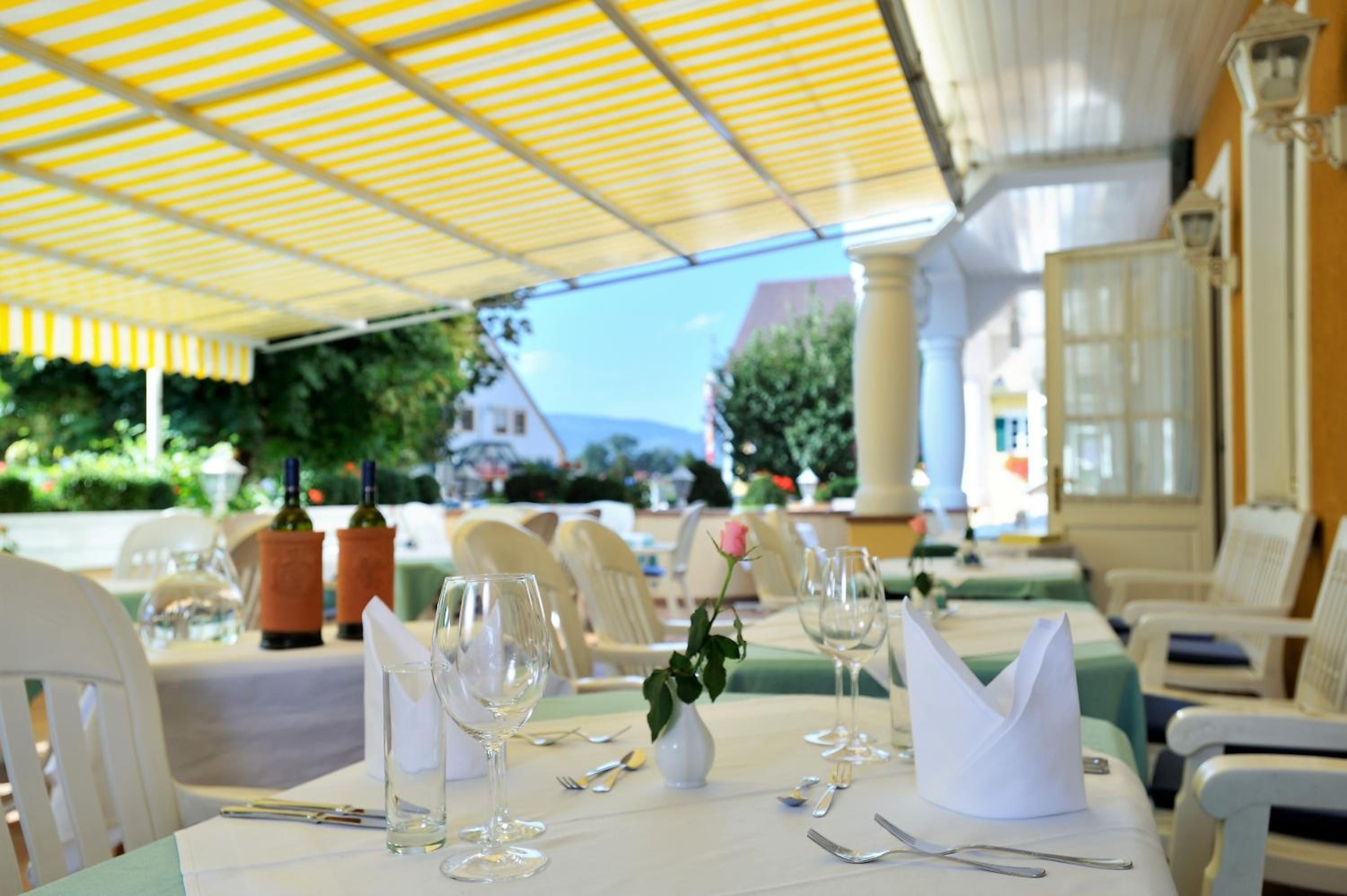 Un área de comedor al aire libre con mesas dispuestas para dos personas, con cristalería transparente y un centro de mesa con rosas.
