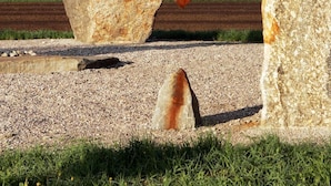 Una gran roca con una escultura metálica que representa a una araña, sobre un paisaje rural con campos y un edificio distante.