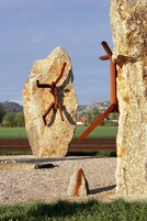 Una gran roca con una escultura metálica que representa a una araña, sobre un paisaje rural con campos y un edificio distante.