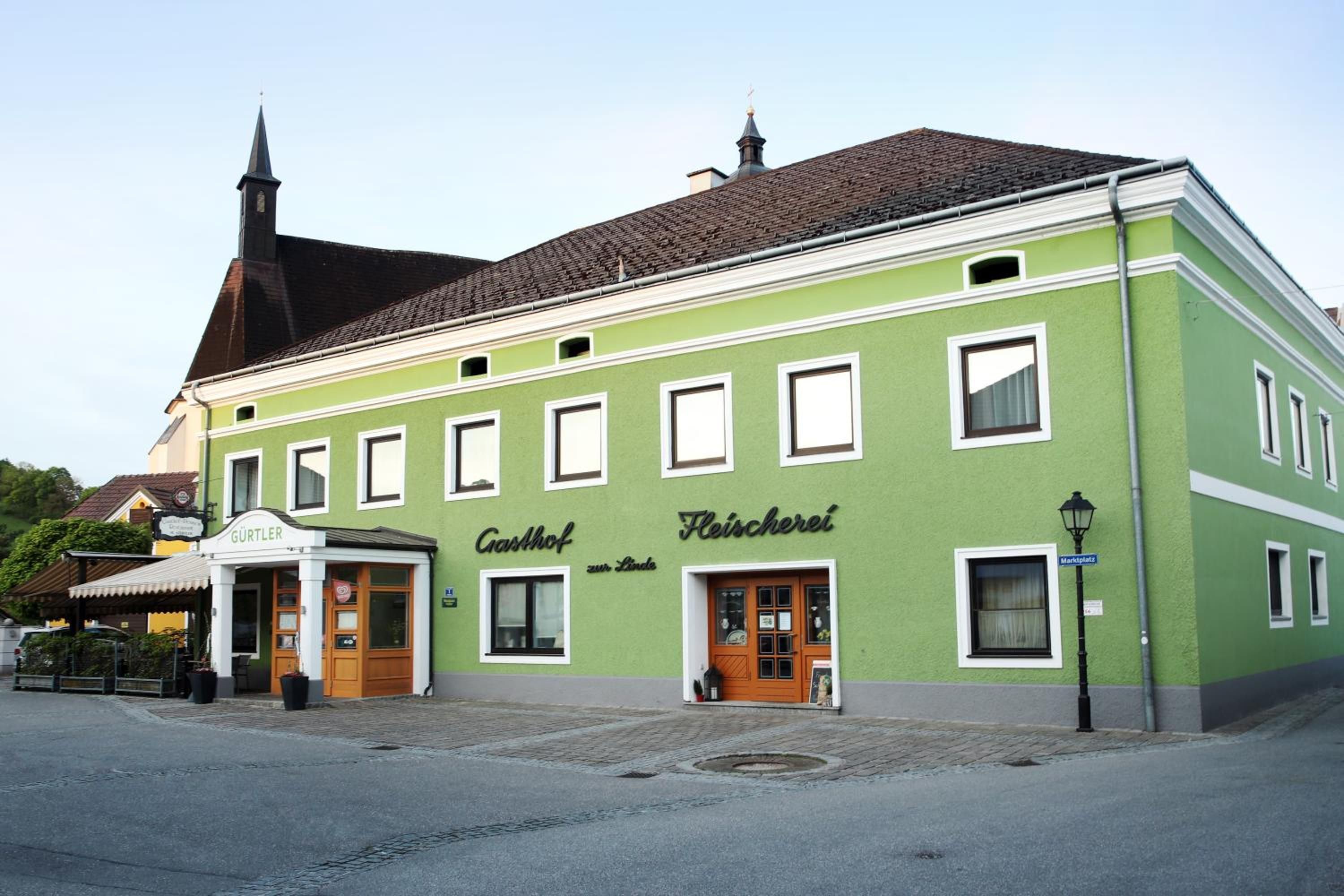 Ein grünes Gebäude mit traditioneller Fassade, im Hintergrund ein Kirchturm und ein Schild mit der Aufschrift 'Gasthof zur Linda'.