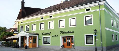 Ein grünes Gebäude mit traditioneller Fassade, im Hintergrund ein Kirchturm und ein Schild mit der Aufschrift 'Gasthof zur Linda'.