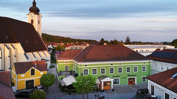 Una plaza de pueblo con una iglesia, un edificio verde con un letrero 'Gürtler' y un edificio amarillo con un letrero 'Jäger'.