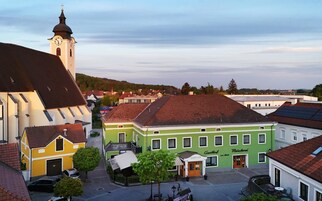 Una plaza de pueblo con una iglesia, un edificio verde con un letrero 'Gürtler' y un edificio amarillo con un letrero 'Jäger'.