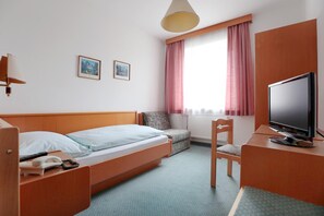 Habitación de hotel con una cama, un escritorio con televisor, una silla, una mesita con teléfono y dos cuadros en la pared.
