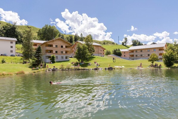 Un hotel frente al lago con varias edificaciones de madera, una zona para nadar y una colina cubierta de césped al fondo.