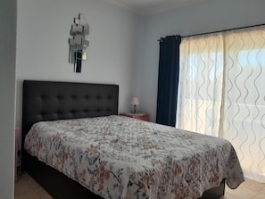 Ein Schlafzimmer mit einem großen Bett, einem Kopfteil, einem Nachttisch mit Lampe und einem Fenster mit Vorhängen.