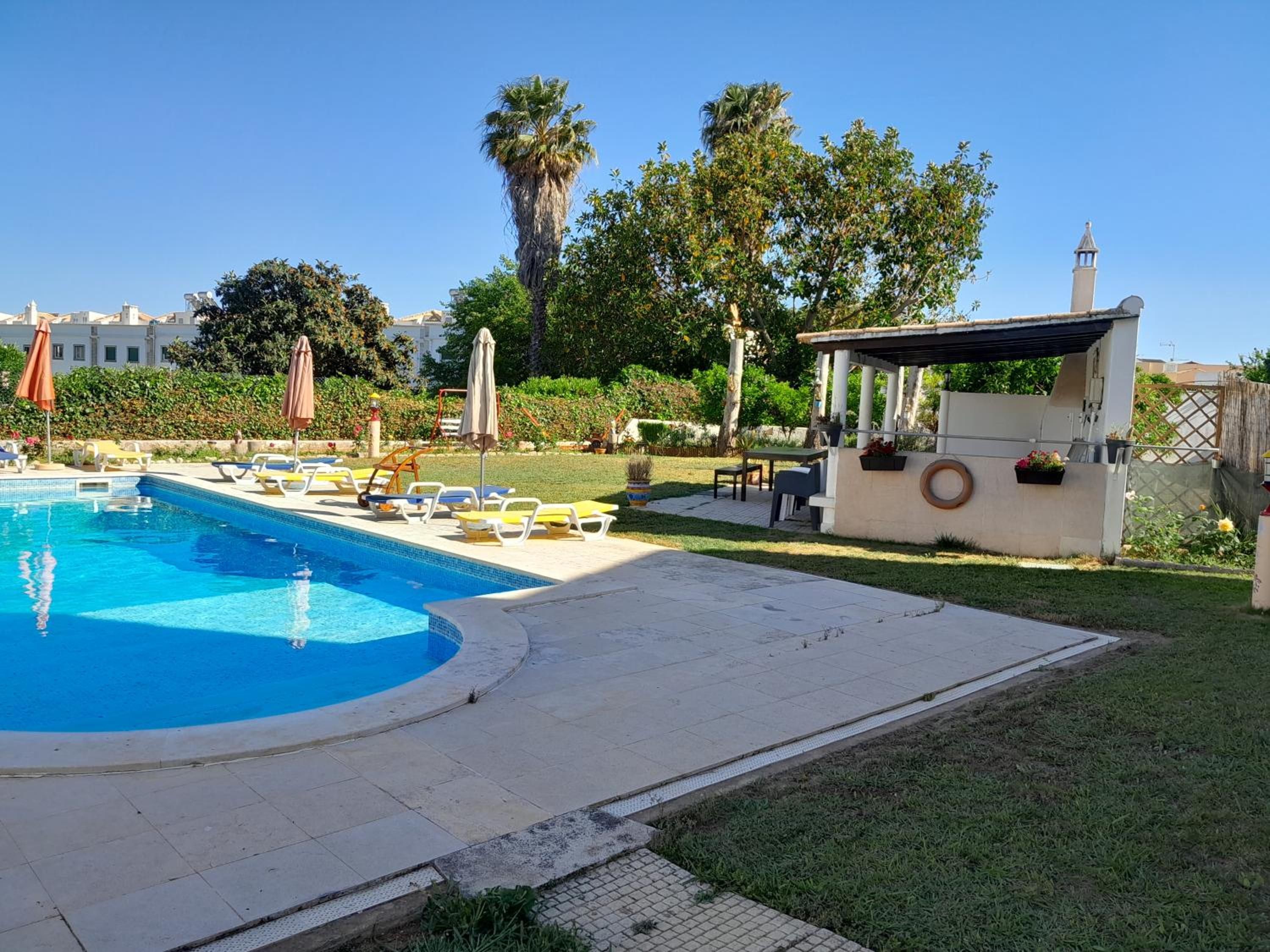 Una piscina con lettini e ombrelloni, una piccola zona relax all'aperto e un giardino con alberi e una recinzione.