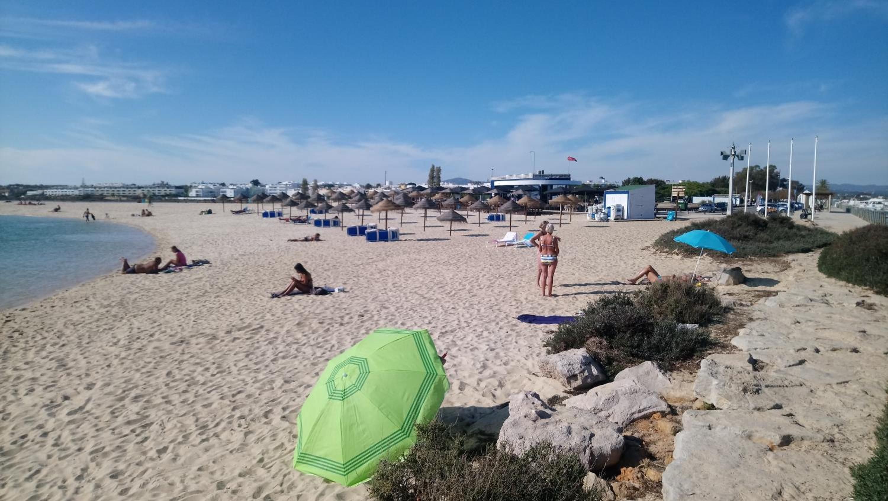A beach with people sunbathing and swimming, umbrellas, and beach huts.