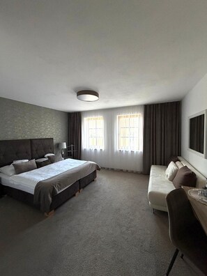 A hotel room with a large bed, a sofa, a desk, and two windows with curtains.