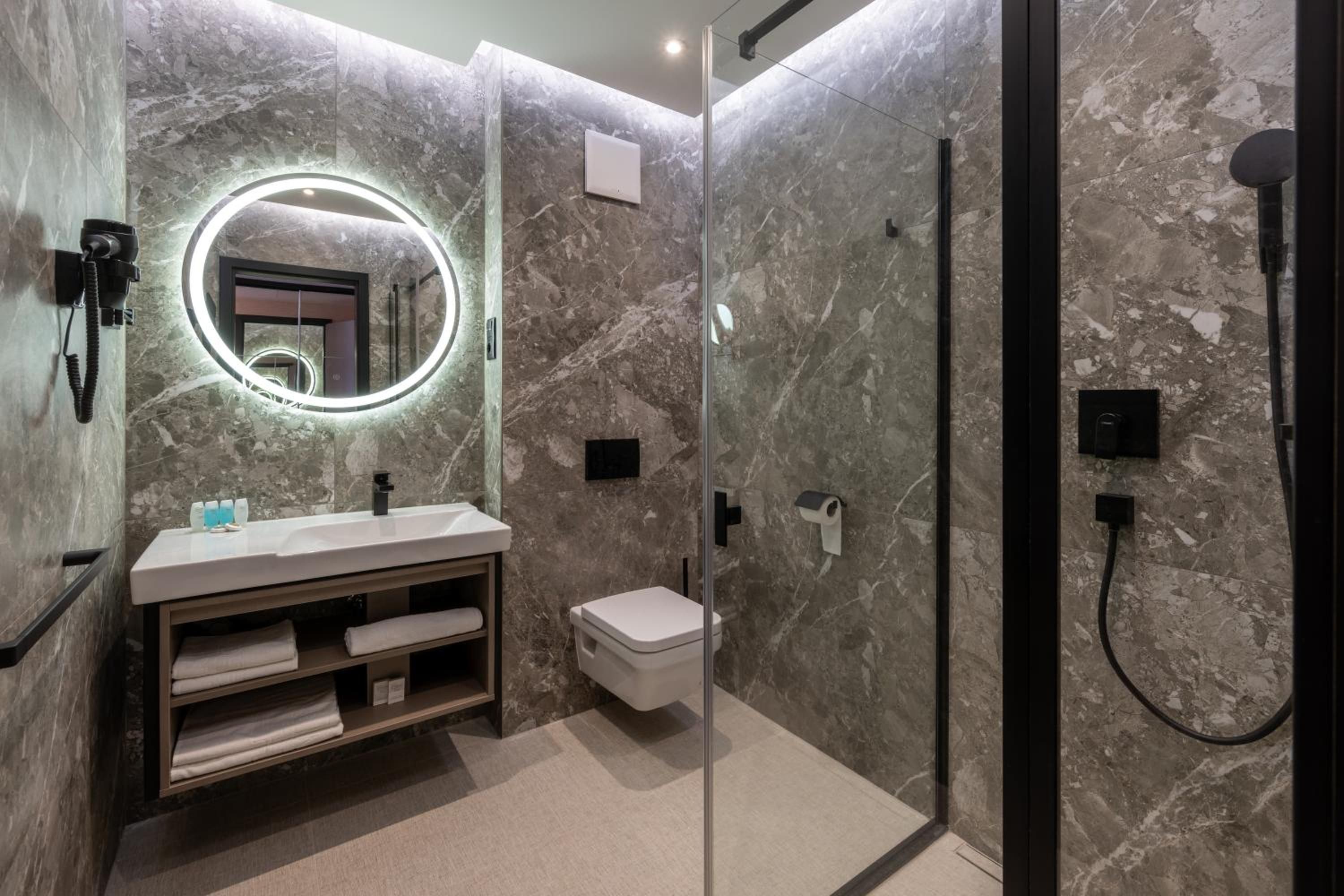 Un baño moderno con pared de mármol, un espejo redondo con iluminación, un lavabo, un inodoro y zona de ducha con mampara de vidrio.