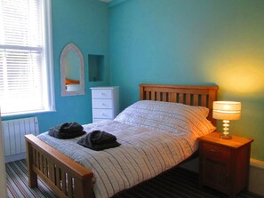 Un dormitorio con cama, una mesita de noche con lámpara, cómoda, espejo y radiador.