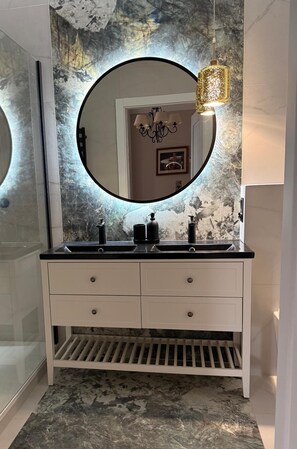 Um banheiro com espelho redondo, duas pias, toucador branco e luminária decorativa.