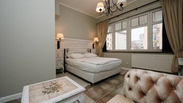 Quarto com cama, abajures, mesa estampada com flores, poltrona com detalhes em capitonê e janela com cortinas.