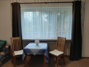 A room with a table and two chairs, a radiator below the window, and a lamp on the table. - Goode Wind (Norden)