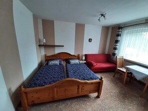 A bedroom with a wooden bed, a red sofa, a small table, and a window with curtains. - Goode Wind (Norden)