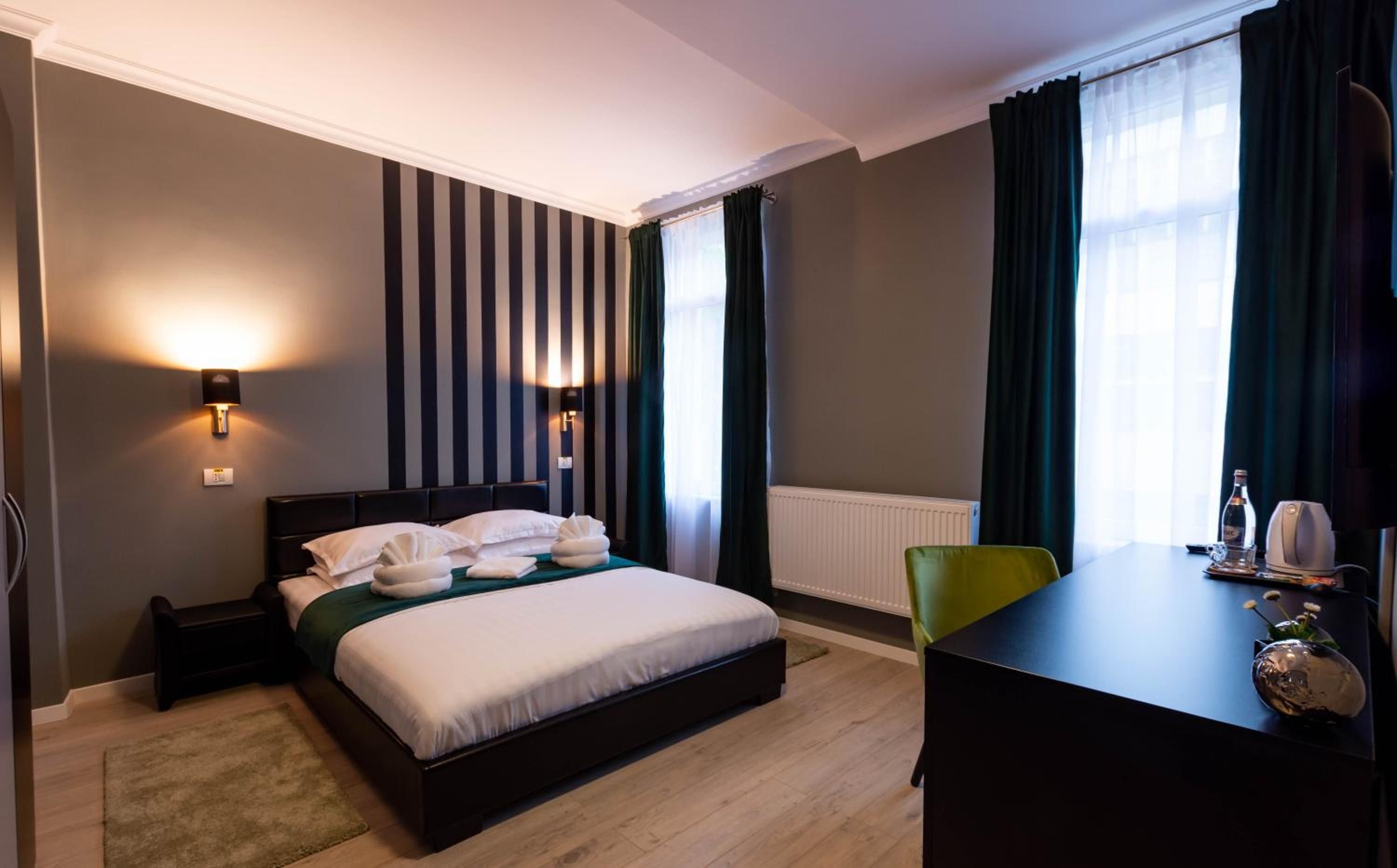 A hotel room with a bed, bedside lamps, a desk, a chair, a radiator, and a window with curtains.
