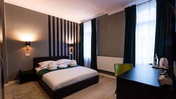A hotel room with a bed, bedside lamps, a desk, a chair, a radiator, and a window with curtains.