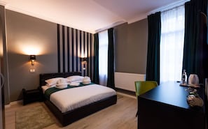 A hotel room with a bed, bedside lamps, a desk, a chair, a radiator, and a window with curtains.