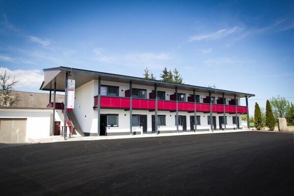 A two-story motel with a flat roof, a covered parking area, and a clear sky. - Burg Rooms 24h Self Check In (Spielberg)