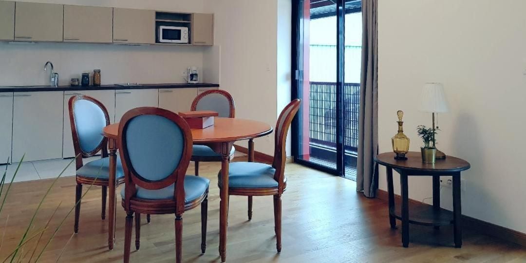 A dining area with a wooden table and chairs, a small side table with a lamp, and a view through a glass door.