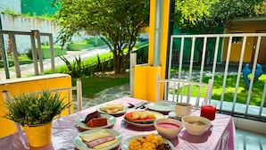 A table set with various dishes, including a slice of cake, a bowl of soup, and a glass of juice, on a balcony with a view of a garden and a building. - Pousada Granville (Campina Grande)
