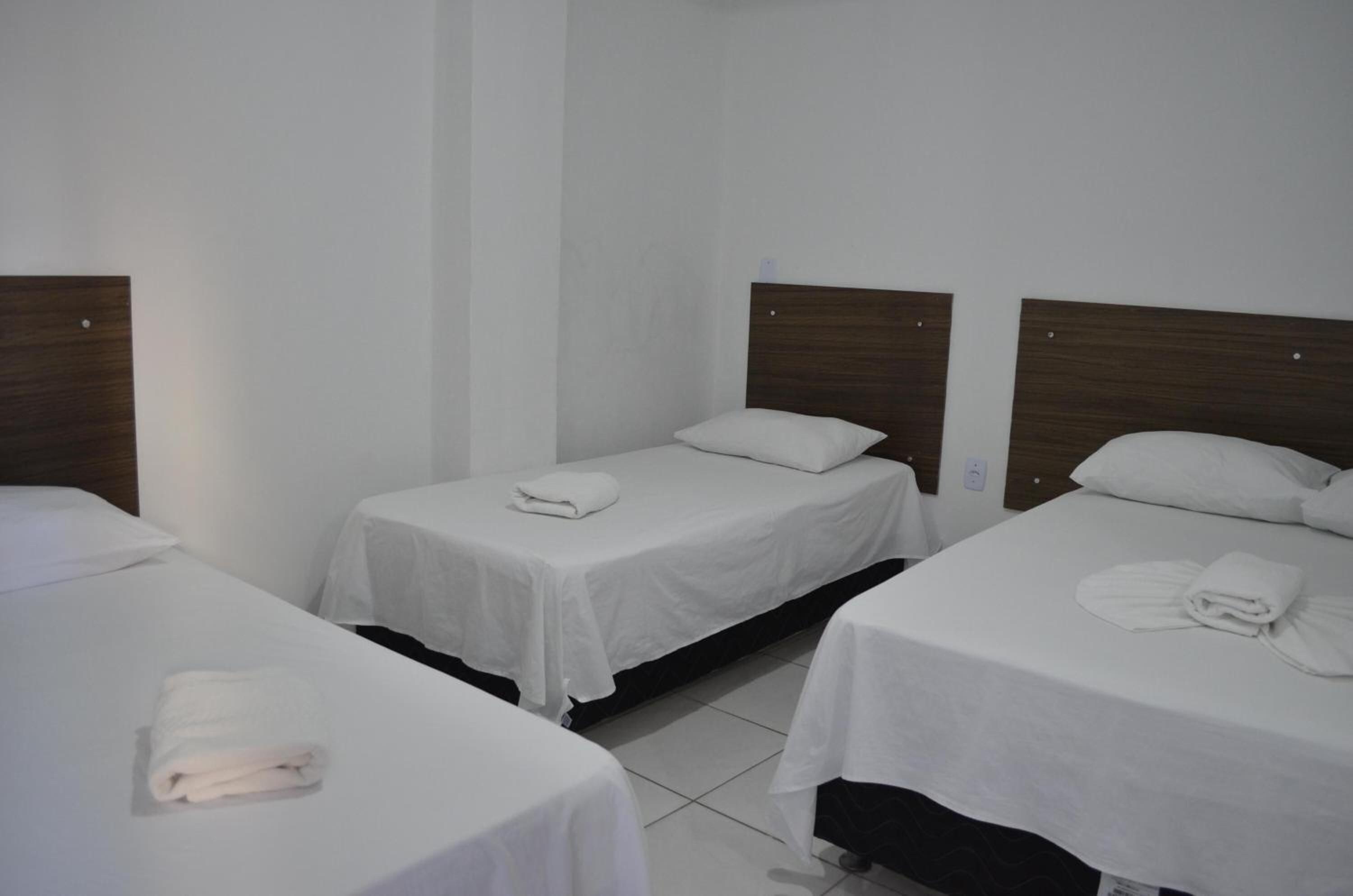 Quarto de hotel com três camas de solteiro, cada uma com uma colcha branca e uma toalha dobrada.