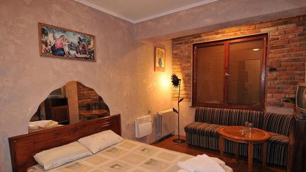 Una habitación de hotel con cama, sofá, una mesita, radiador, ventana con persianas y un cuadro en la pared.