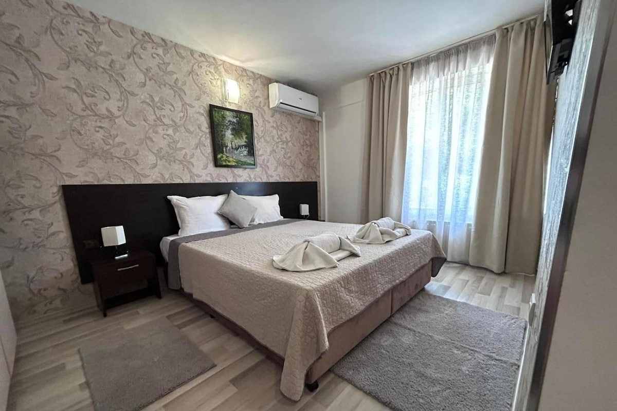 Una habitación de hotel con una cama grande, una mesita de noche, un equipo de aire acondicionado, un cuadro en la pared y una ventana con cortinas.
