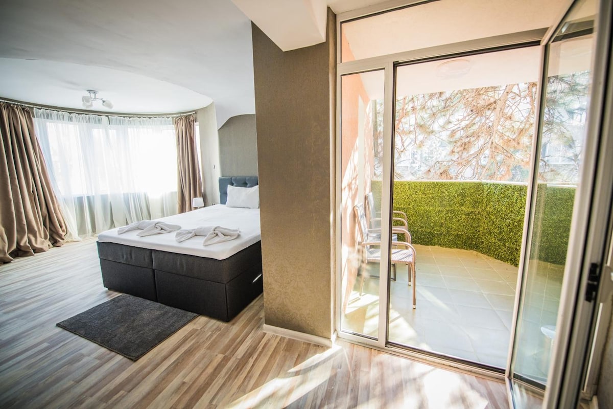 A modern bedroom with a large bed, a sliding glass door to a balcony, and a view of a hedge.