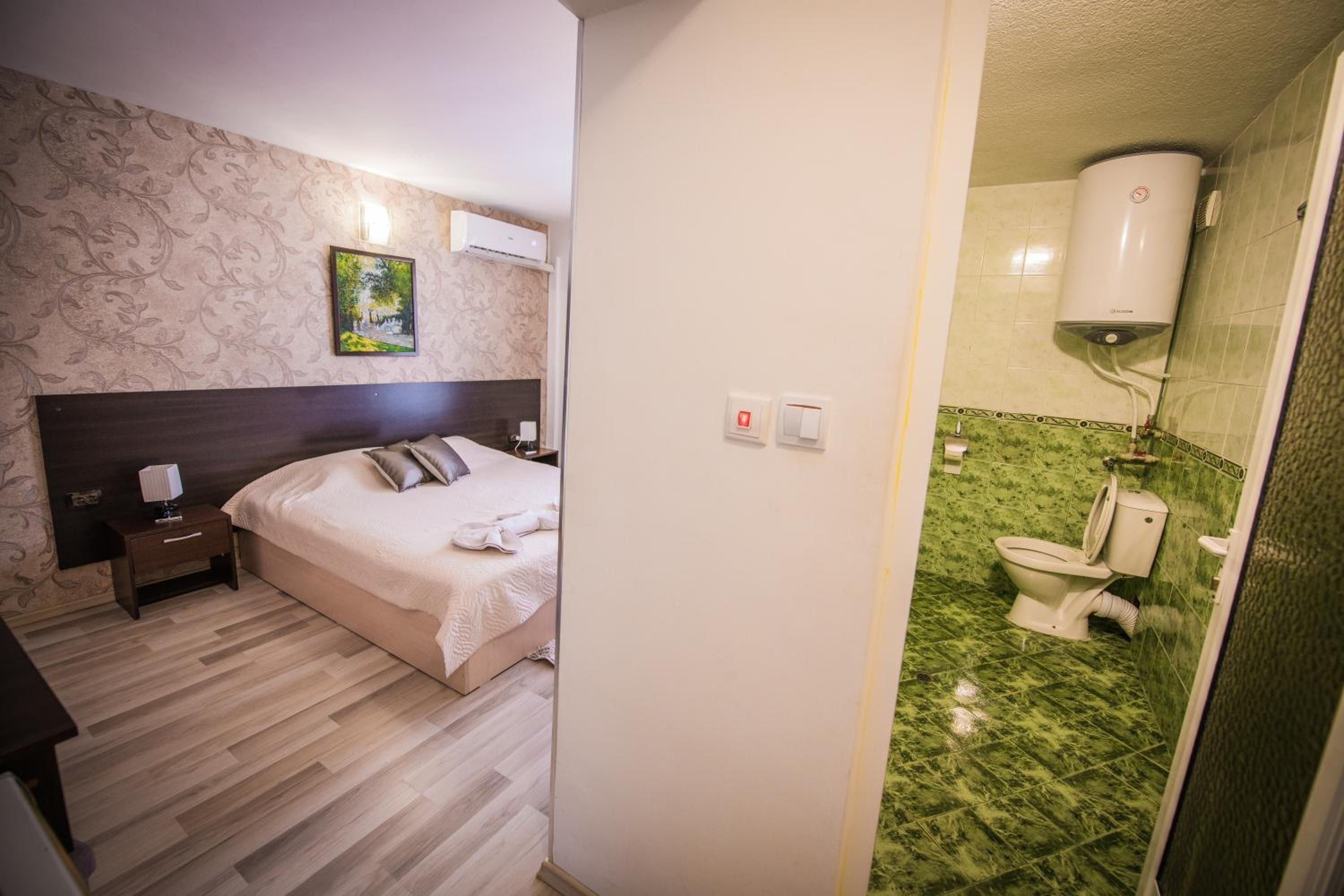 Habitación de hotel con cama, mesita de noche, lámpara de noche, equipo de aire acondicionado, un cuadro en la pared y un baño con inodoro y paredes de azulejos verdes.