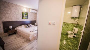 Habitación de hotel con cama, mesita de noche, lámpara de noche, equipo de aire acondicionado, un cuadro en la pared y un baño con inodoro y paredes de azulejos verdes.