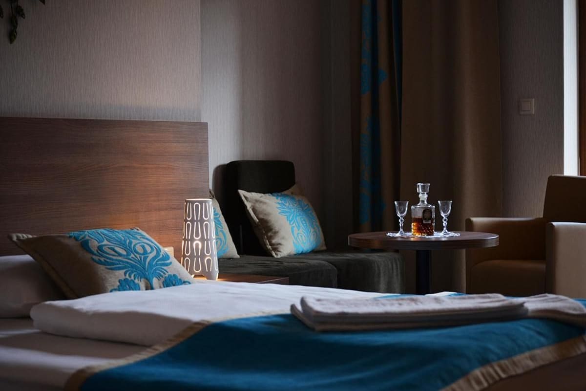Una habitación de hotel con una cama, una silla, una mesita pequeña con vasos y una botella, y una mesita de noche.