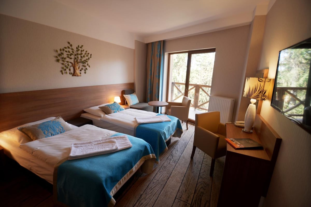 Habitación de hotel con dos camas, un escritorio, una silla, una lámpara y ventana con vista a árboles.