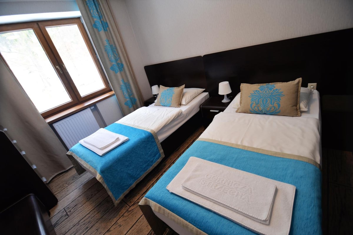 Habitación de hotel con dos camas, cada una con cubrecama azul y ropa blanca, cabeceros oscuros y una ventana con cortinas.
