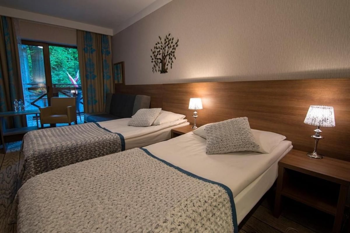 Habitación de hotel con dos camas, un cabecero de madera, una mesita de noche con lámpara y vistas a un balcón con cortinas.