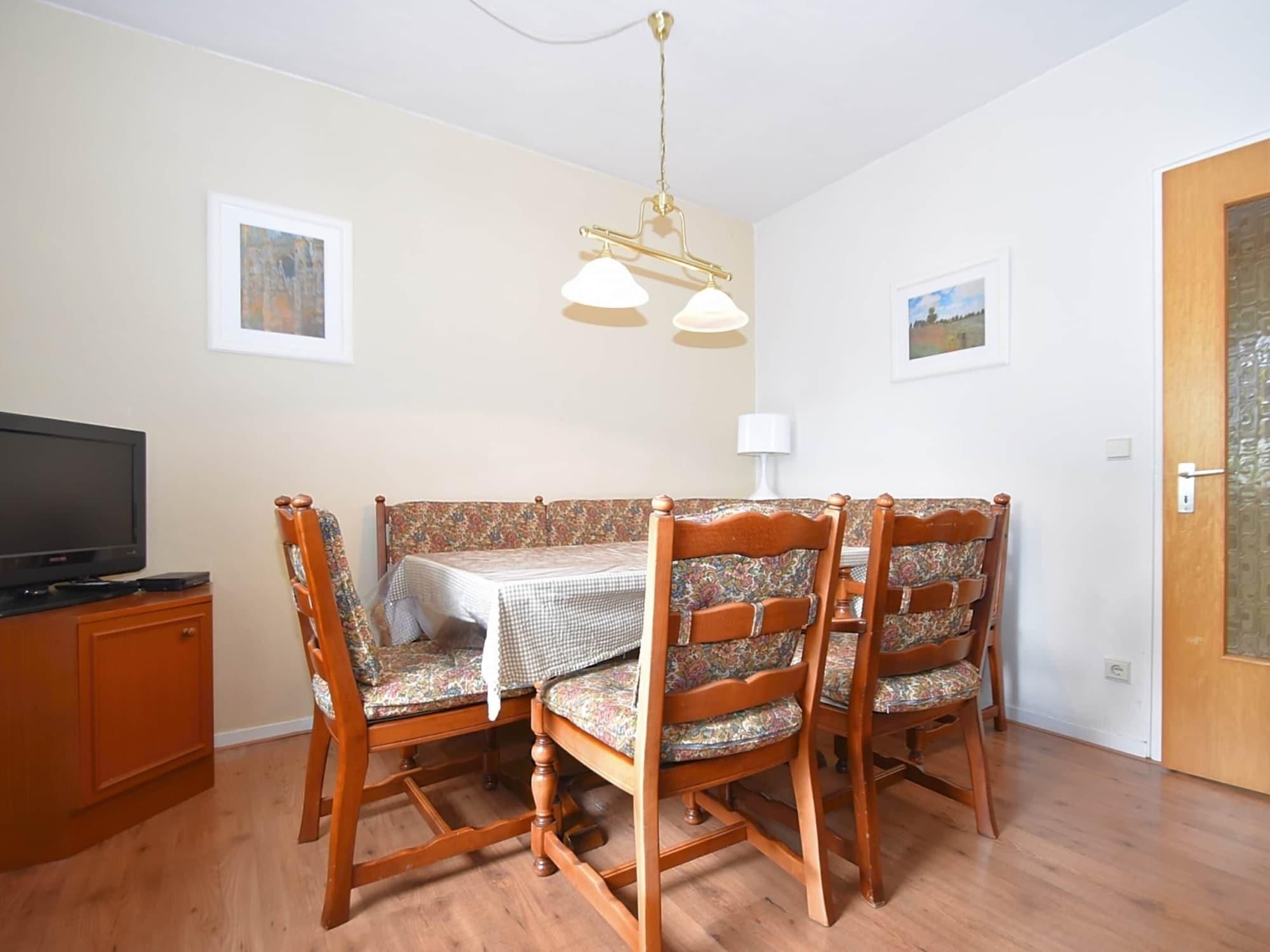 A dining area with a table set for four, wooden chairs with patterned upholstery, a television on a cabinet, and two wall-mounted lamps.