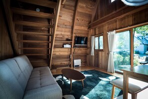 A cozy cabin interior with a sofa, a dining table, a TV, and a large window offering a view of the outdoors. - Yukiita Lodge (Hakuba)