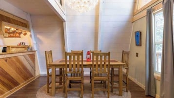 Uma área de refeições com mesa e cadeiras de madeira, um lustre e vista para o exterior através de uma janela.